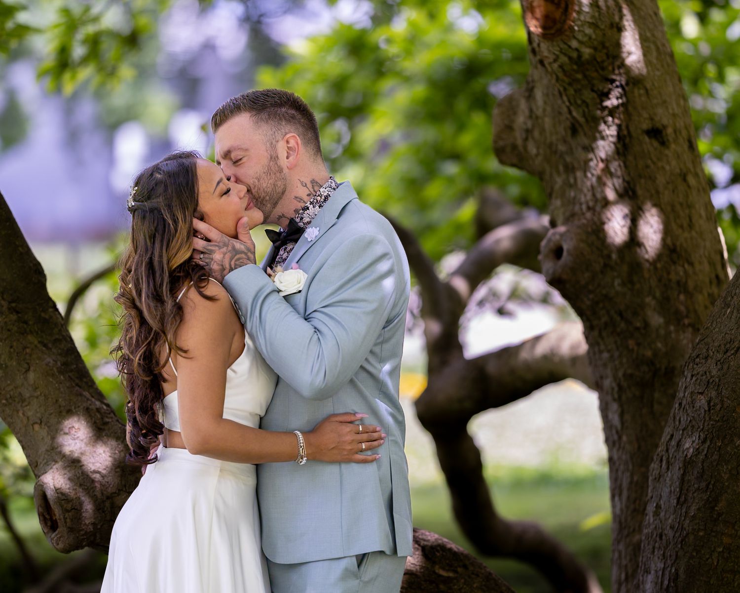 Couple kissing outdoors under a tree in natural setting, wearing casual formal attire.