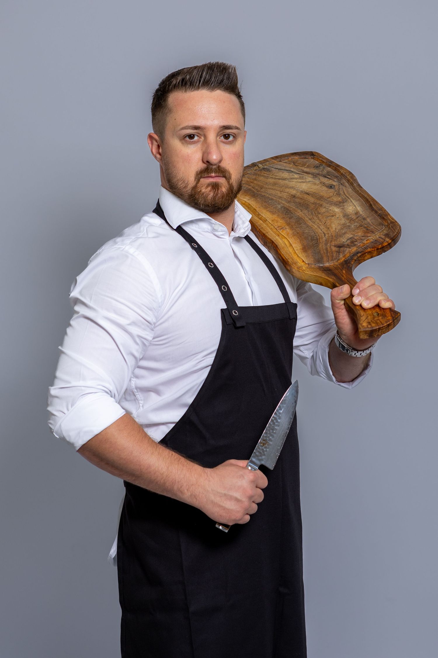 A male chef in a black apron holds a large knife and rustic wooden cutting board against a gray background.