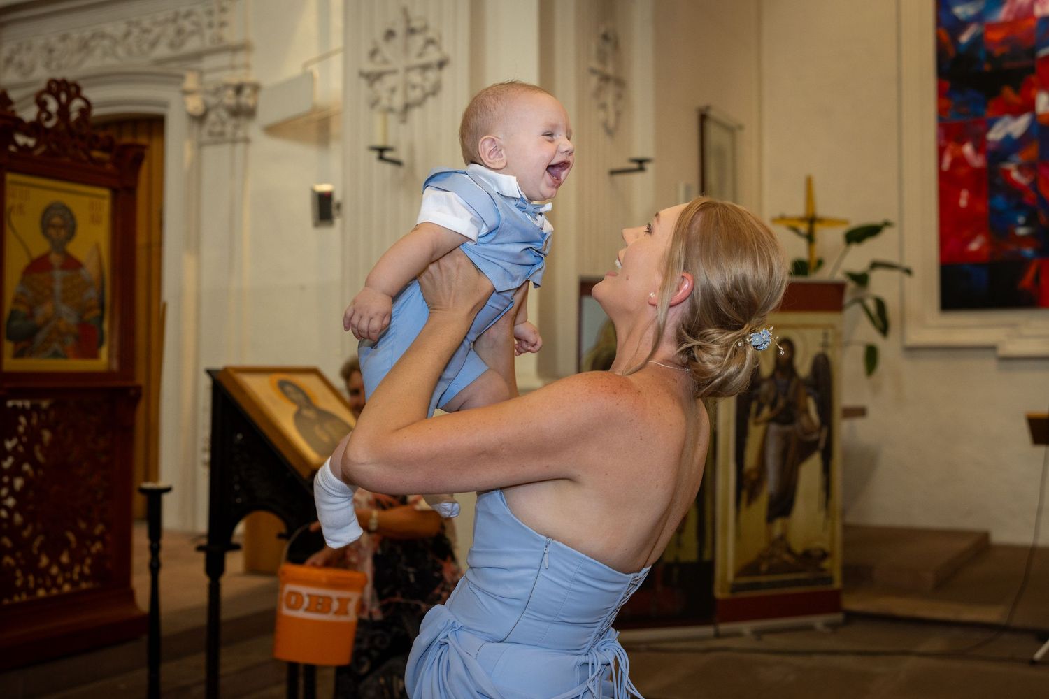 Eine Frau im hellblauen Kleid hebt den lachenden Aris in der Kirche hoch.