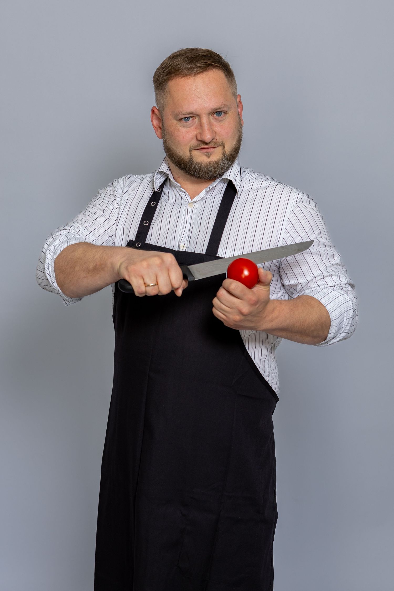 Bearded male chef in black apron holding a knife and red tomato against a gray background.