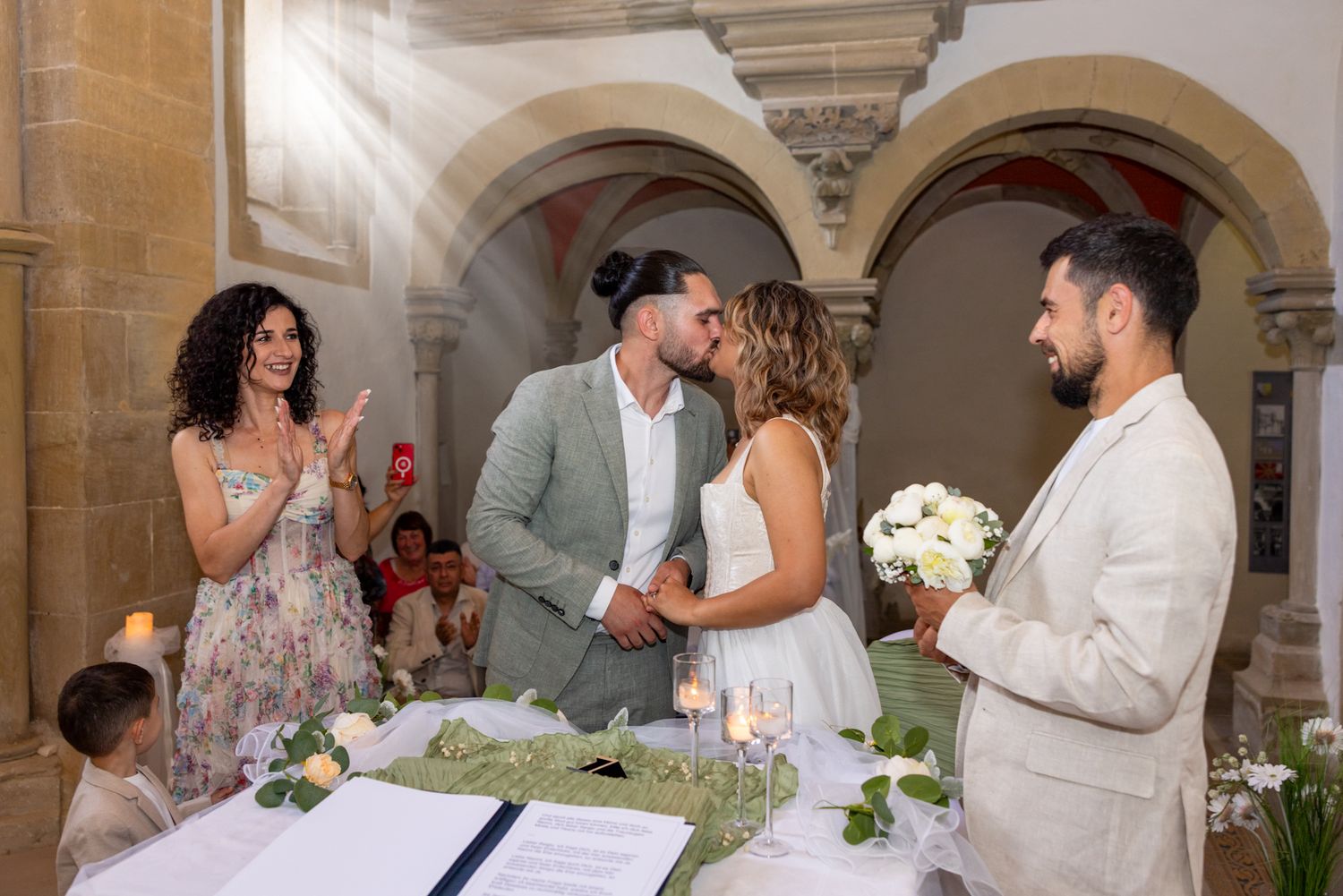 Couple shares a kiss at wedding ceremony while guests applaud in elegant venue.