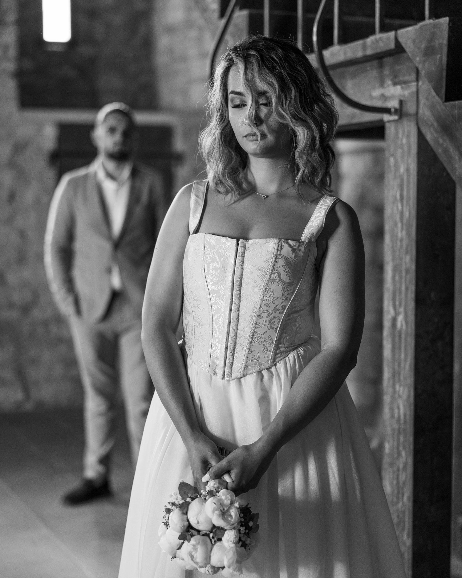 Bride in white dress holding bouquet with groom standing in background in black and white wedding photo.
