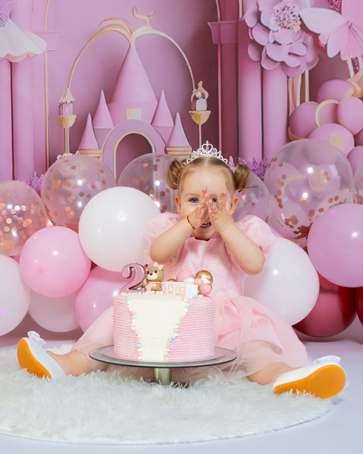A toddler in a pink dress and tiara celebrates her 2nd birthday with a cake, surrounded by pink balloons and a castle backdrop.