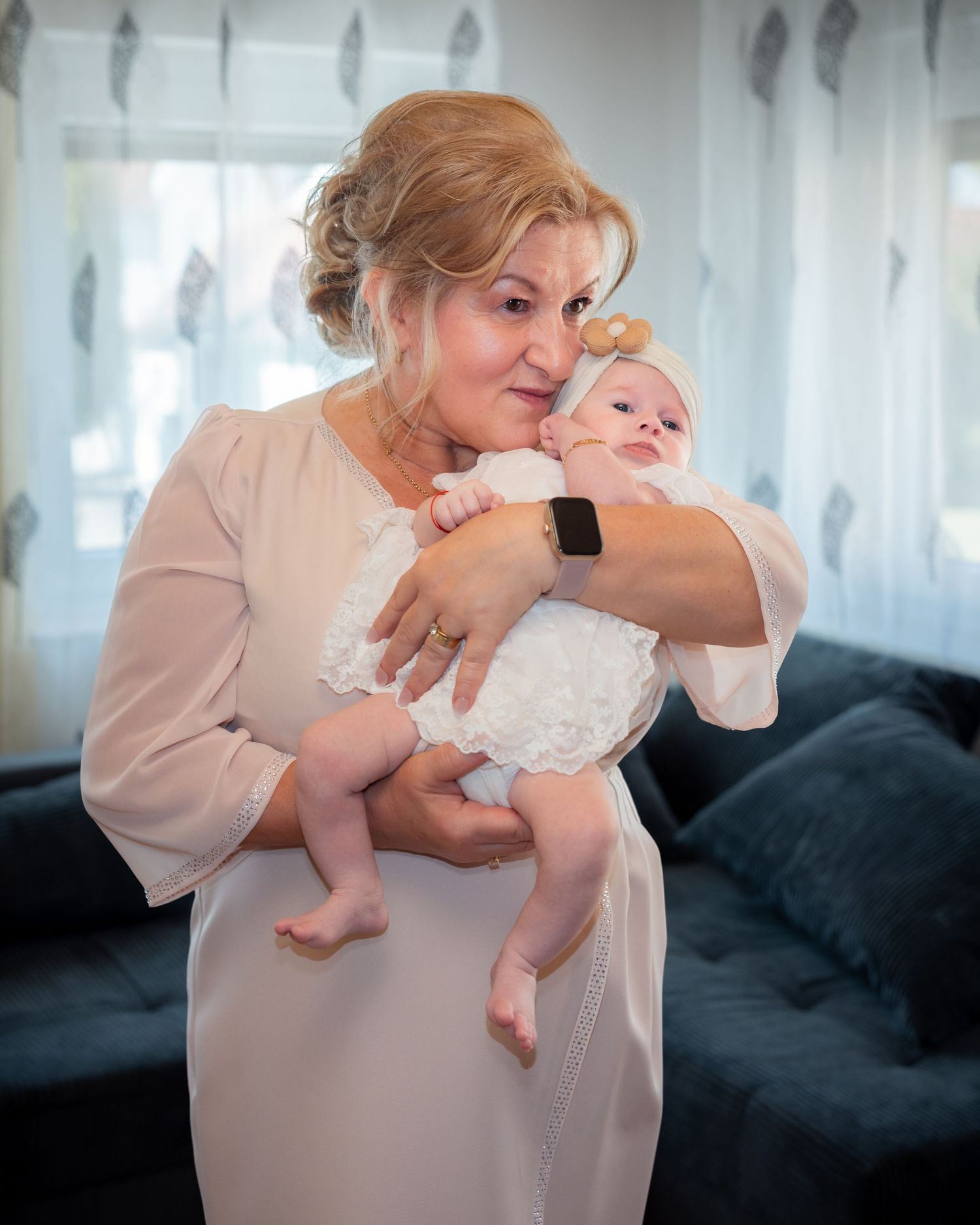 Laras Großmutter hält sie im weißen Taufkleid auf dem Arm vor hellen Vorhängen.