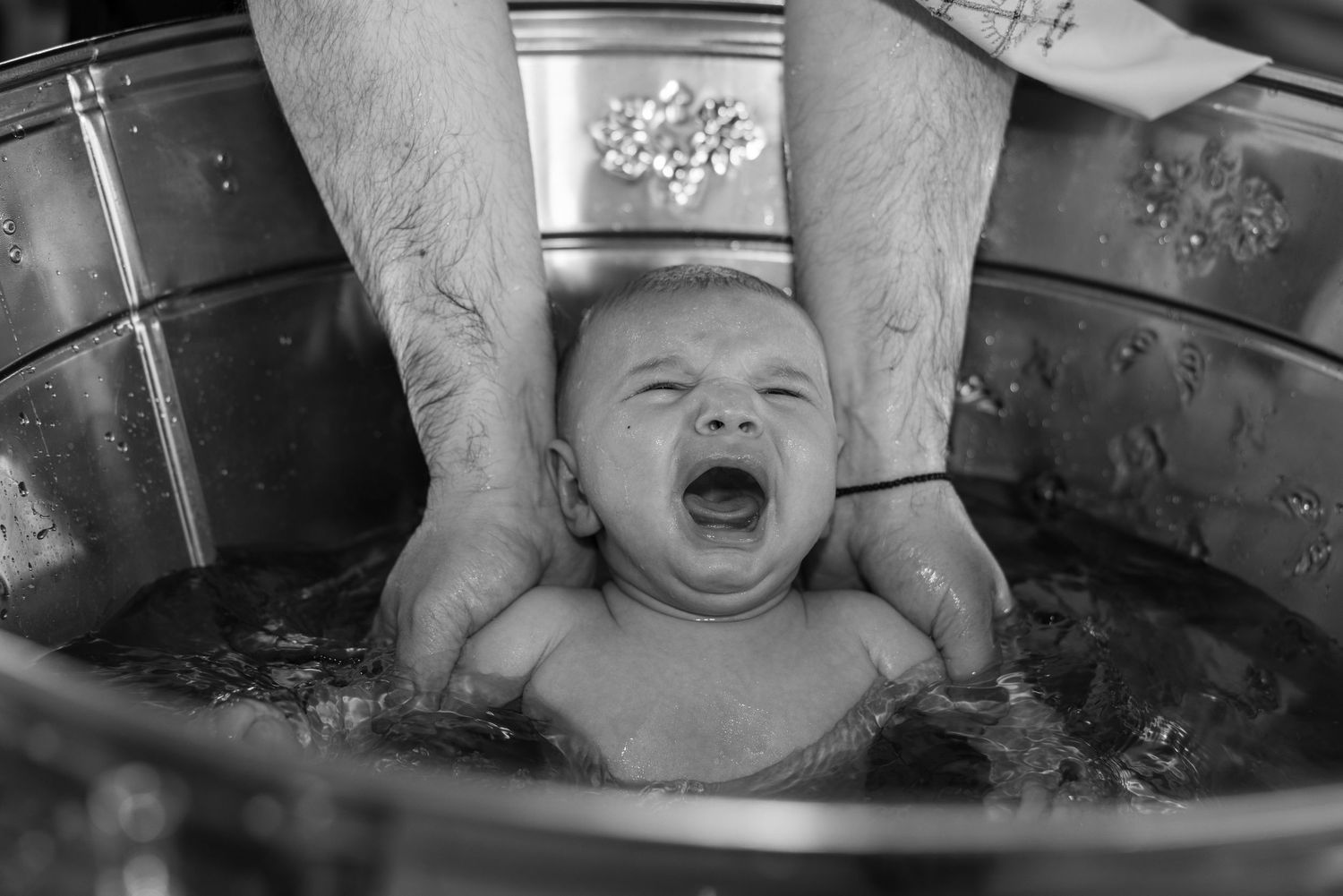 Black and white photo of a crying newborn baby being baptized in a traditional christening font.