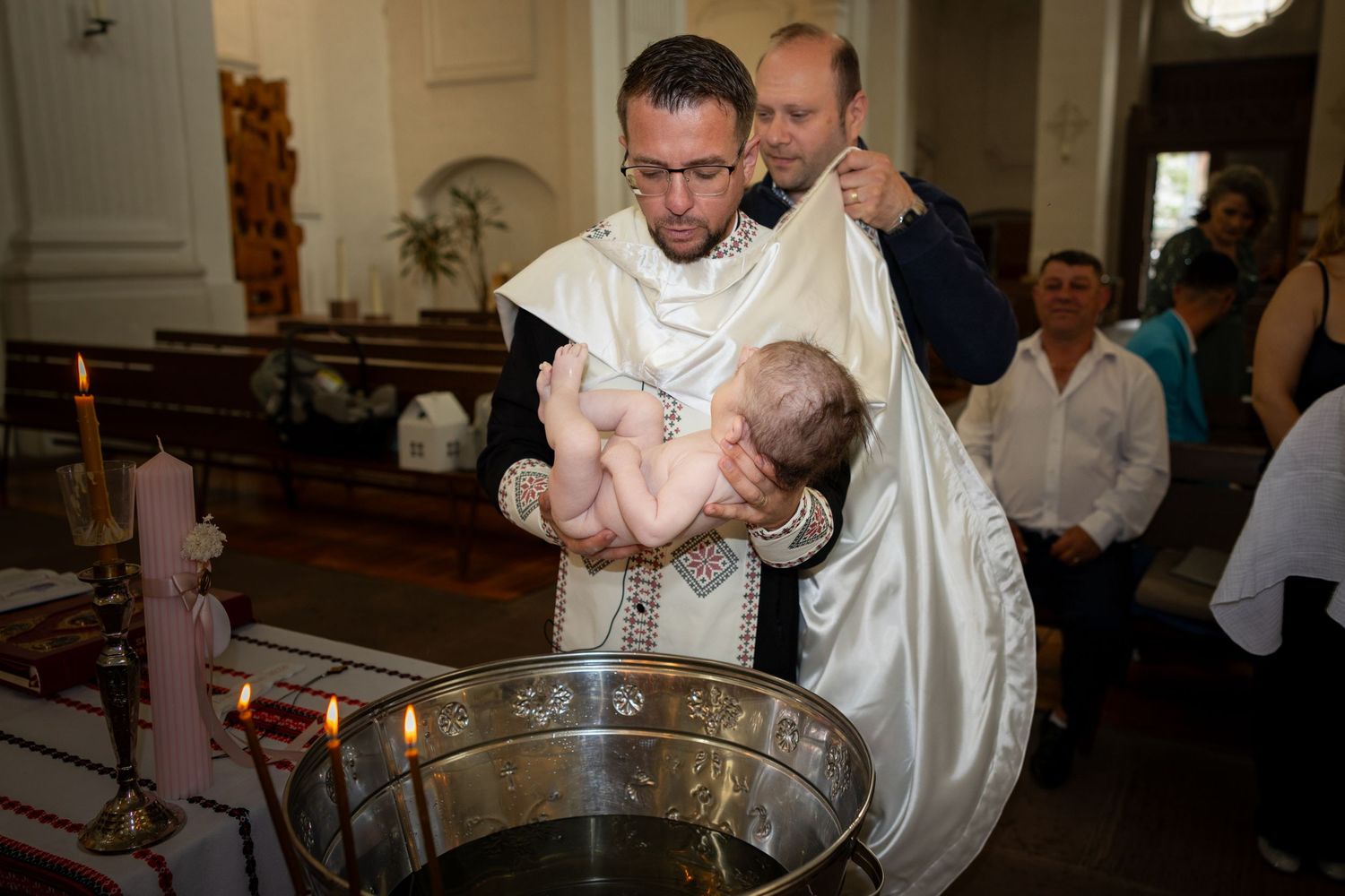Der Priester hält Alexia Ioana über dem Taufbecken, während ein Mann hinter ihm ein weißes Tuch anhebt.