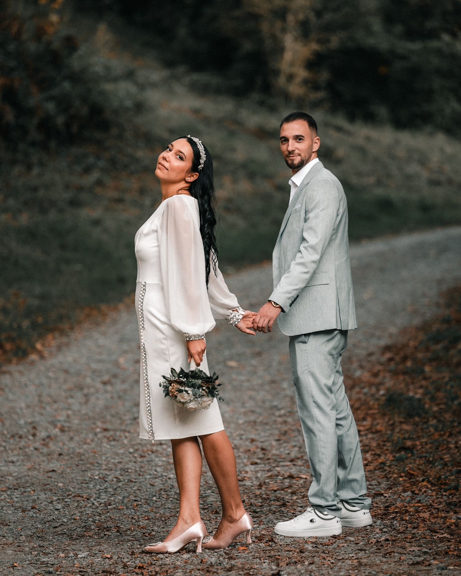 Bride and groom holding hands on a rural road, dressed in casual wedding attire with a natural outdoor setting.