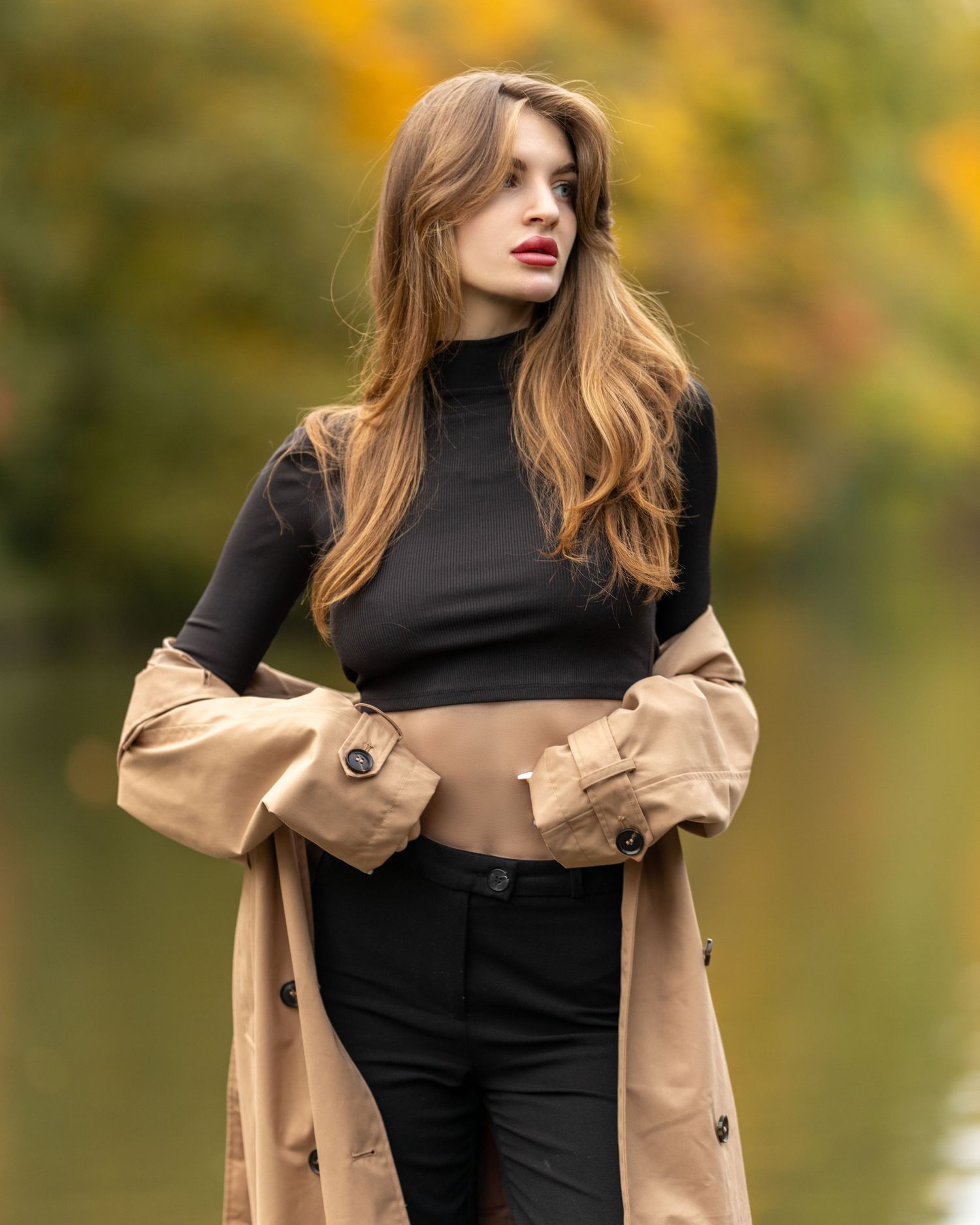 Young woman in black turtleneck and beige trench coat posing outdoors near a lake with autumn foliage background.