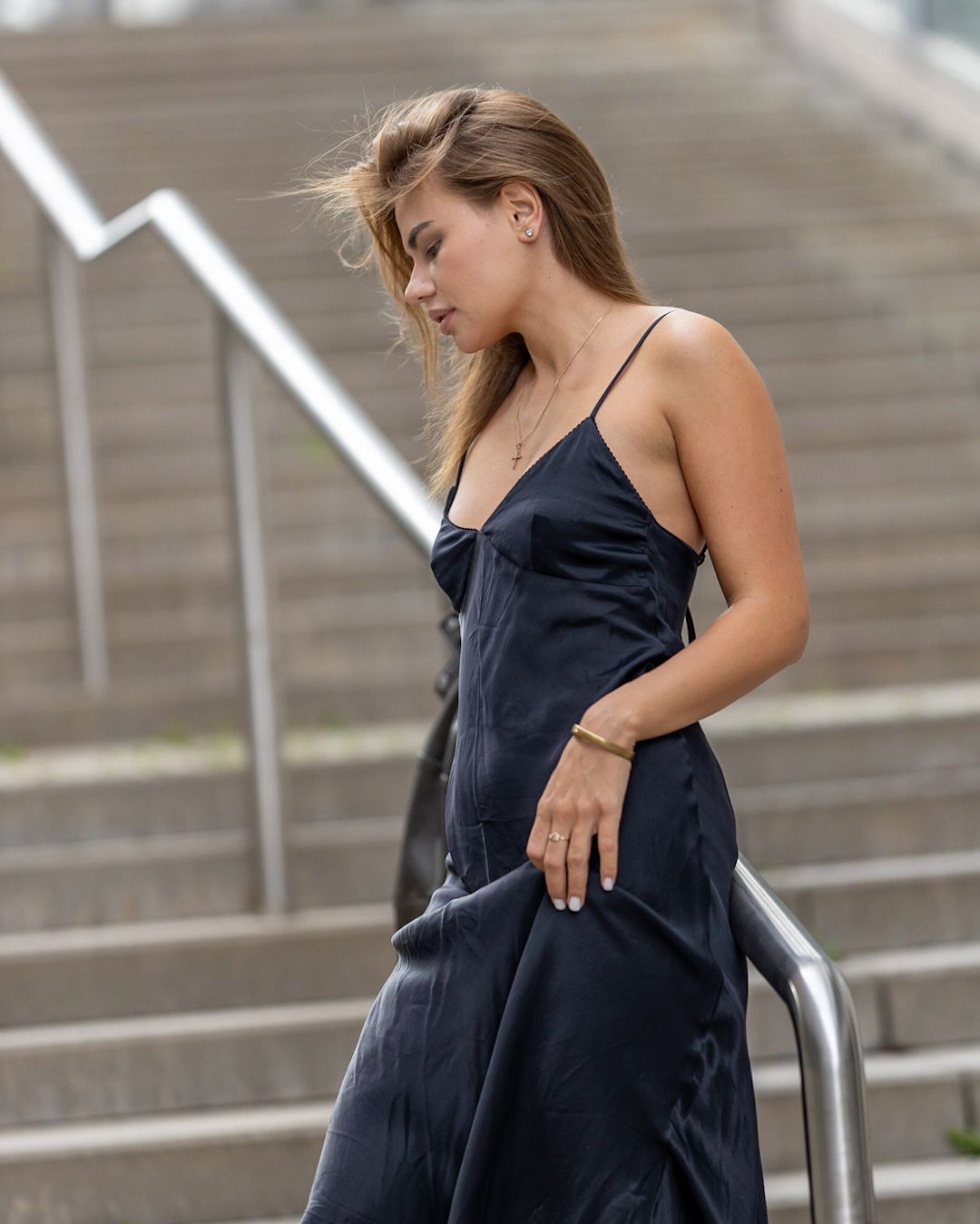 Young woman in elegant black satin slip dress posing on urban staircase with wind-blown hair.
