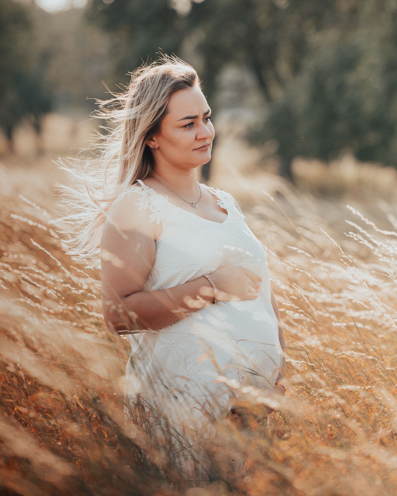 Pregnant woman in white dress standing in golden field with wind-blown hair in warm sunlight.