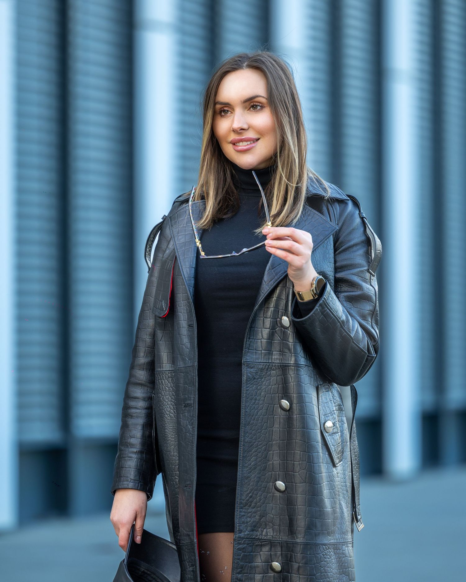 Woman in black leather trench coat and dress holding sunglasses, posing outdoors with blurred blue background.