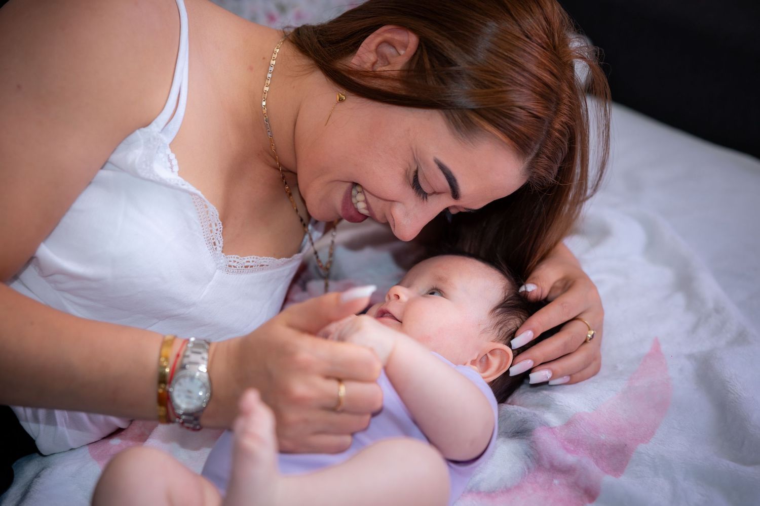 A smiling mother tenderly gazes at her newborn baby while lying together on a soft white bed.