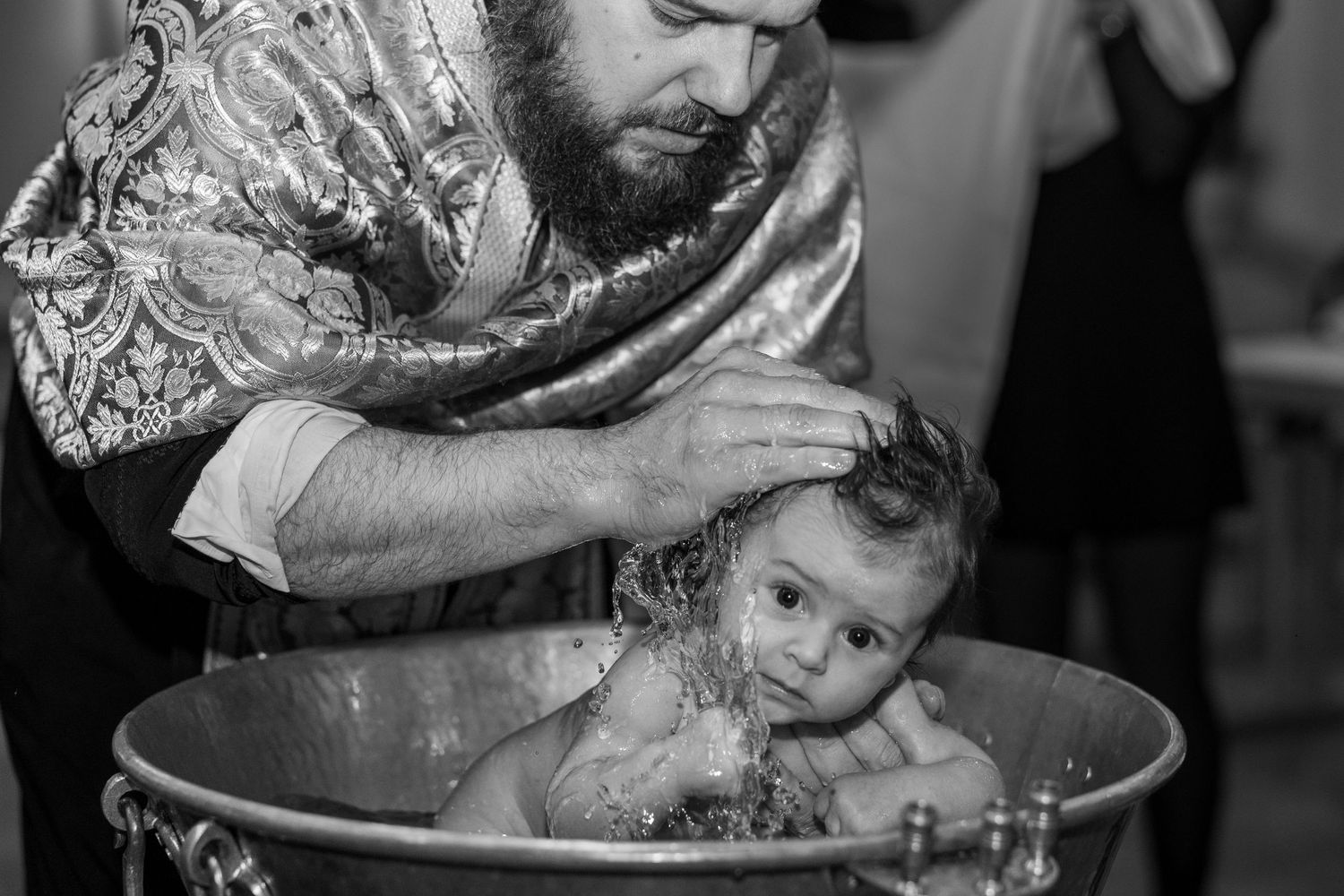 Black and white photo of a priest performing a baby baptism in a traditional metal font.