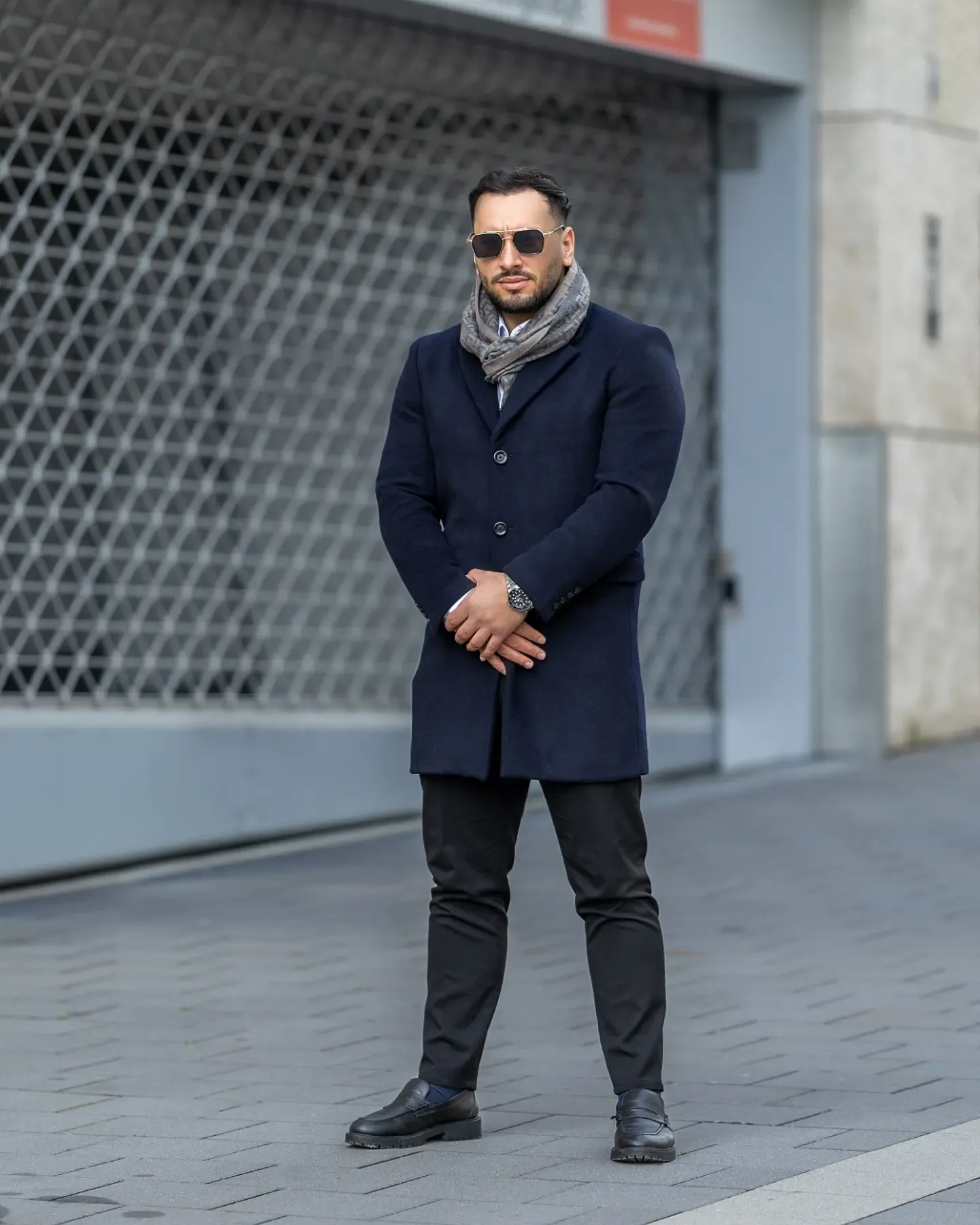 Man in navy coat, gray scarf, sunglasses, black pants and boots poses outdoors on cobblestone street.