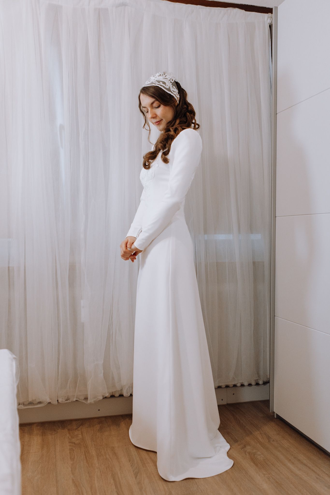 Bride in elegant long-sleeve white wedding gown with tiara, posing in a bright dressing room.