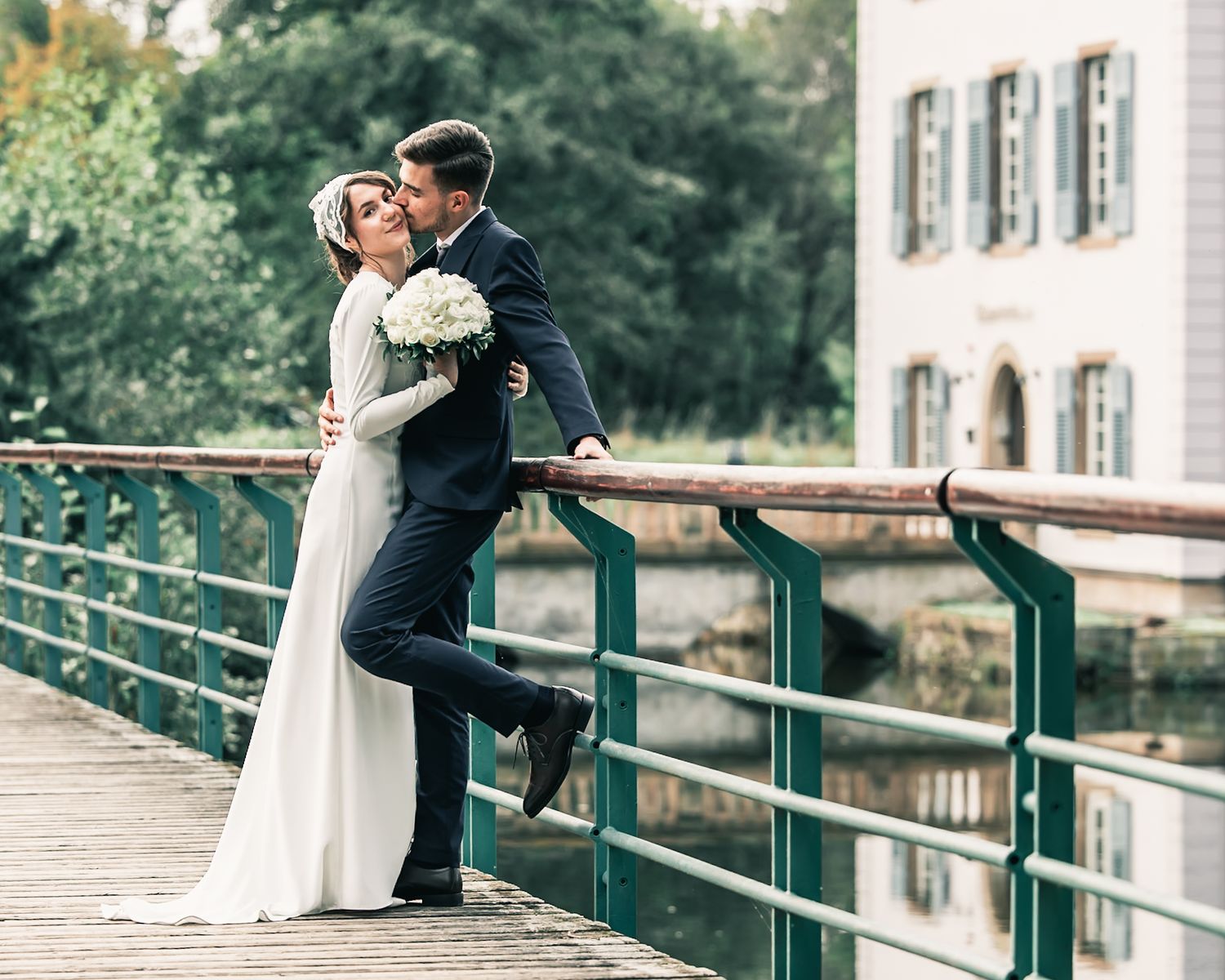 A couple in wedding attire shares a romantic kiss on a bridge, with lush greenery and a historic building in the background.