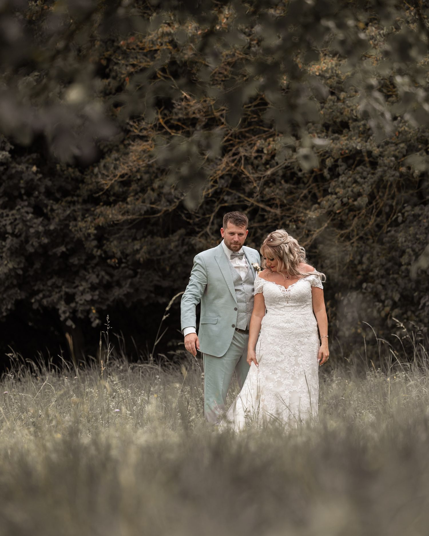 Bride and groom walking through a meadow in front of lush trees, wearing lace wedding dress and sage suit.