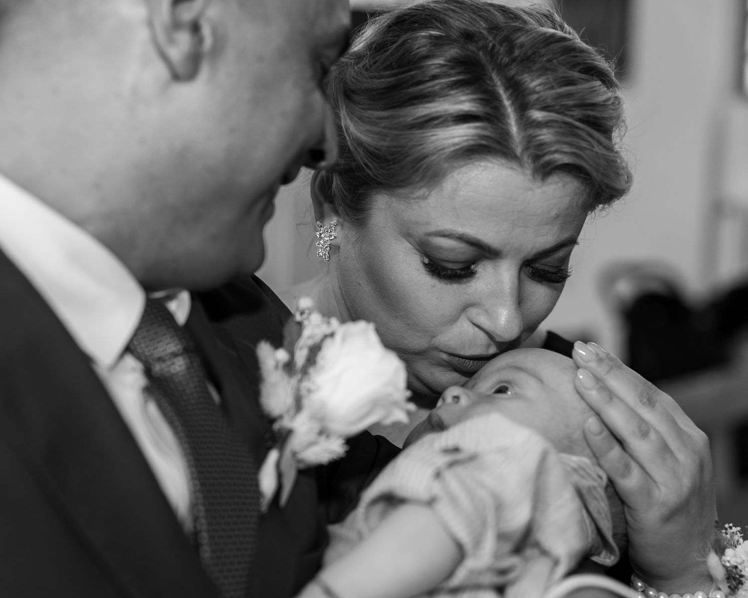 Black and white photo of a bride and groom tenderly kissing a newborn baby at their wedding ceremony.