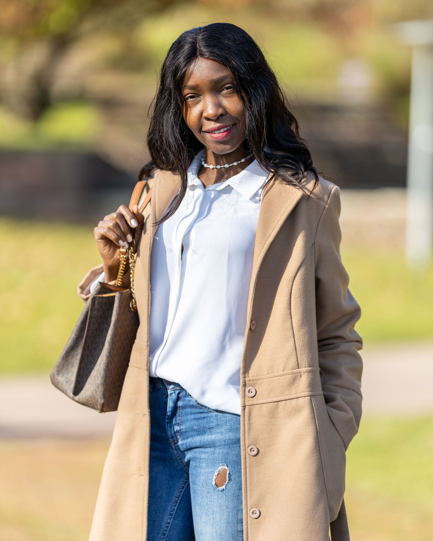 Jacky posiert in hellem Mantel und Jeans mit einer Handtasche im Park