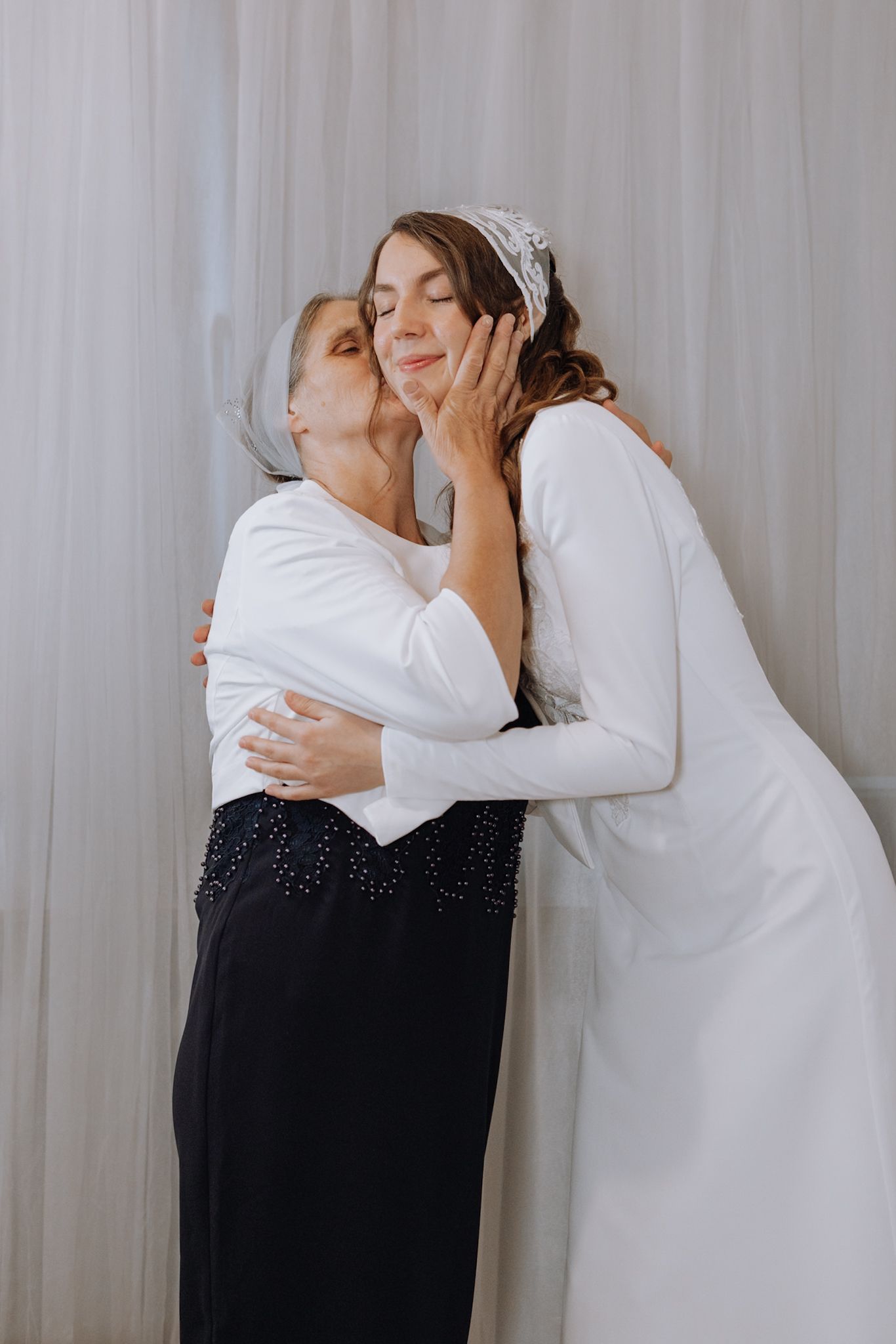 A bride in white dress and headpiece shares a tender embrace and kiss with an older woman in black dress against sheer curtains.
