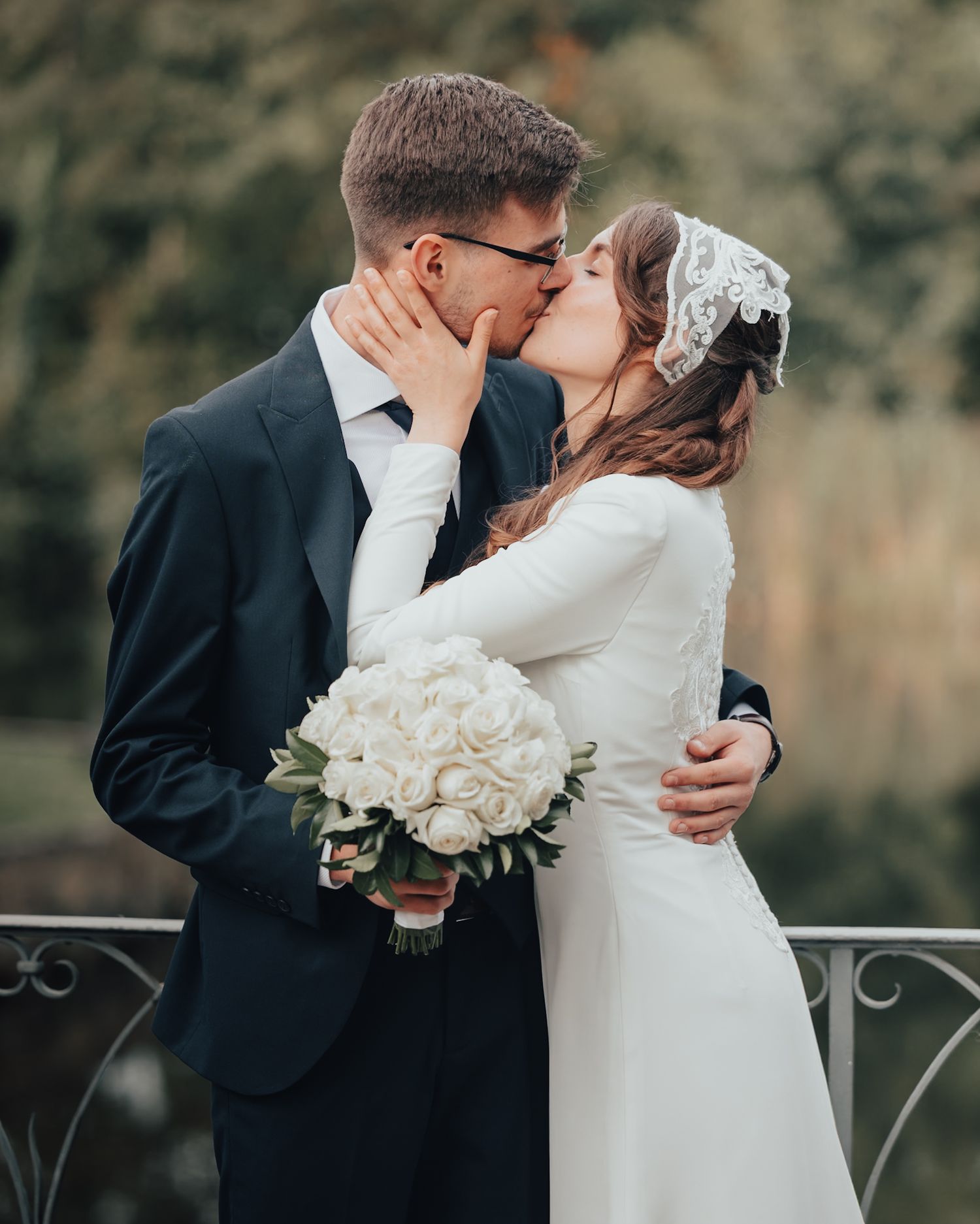 Bride and groom kissing outdoors while holding white wedding bouquet, wearing formal attire with veil and suit.