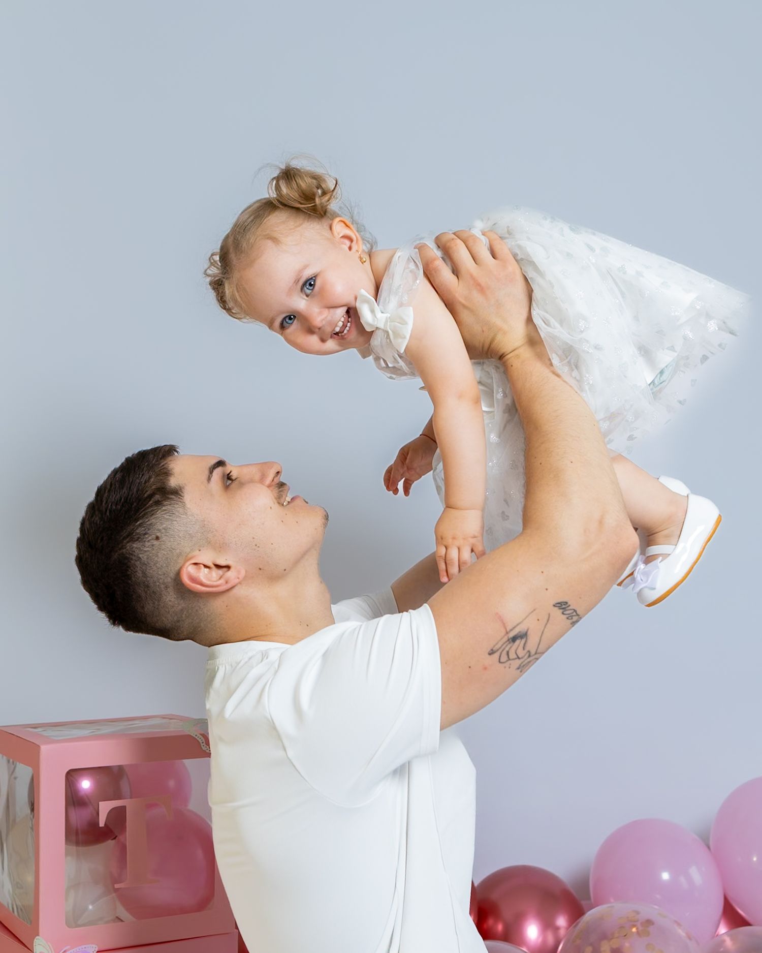 Father in white shirt lifting and playing with happy toddler daughter in white dress against light blue background.