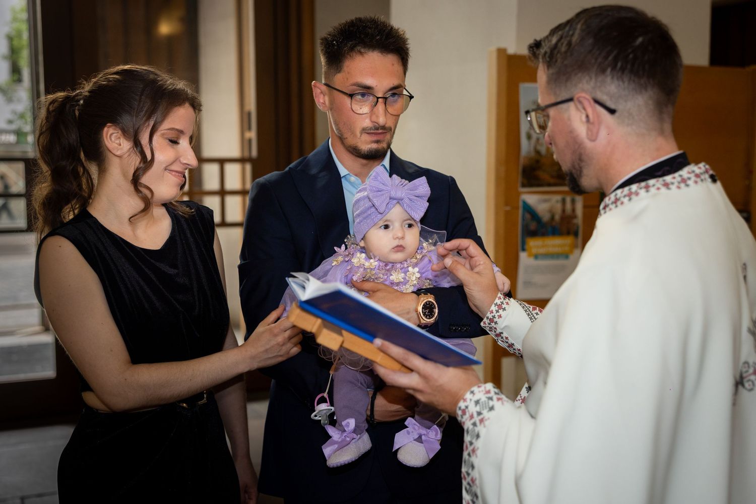 Parents present baby in purple outfit to priest during a Christian baptism ceremony in a church.