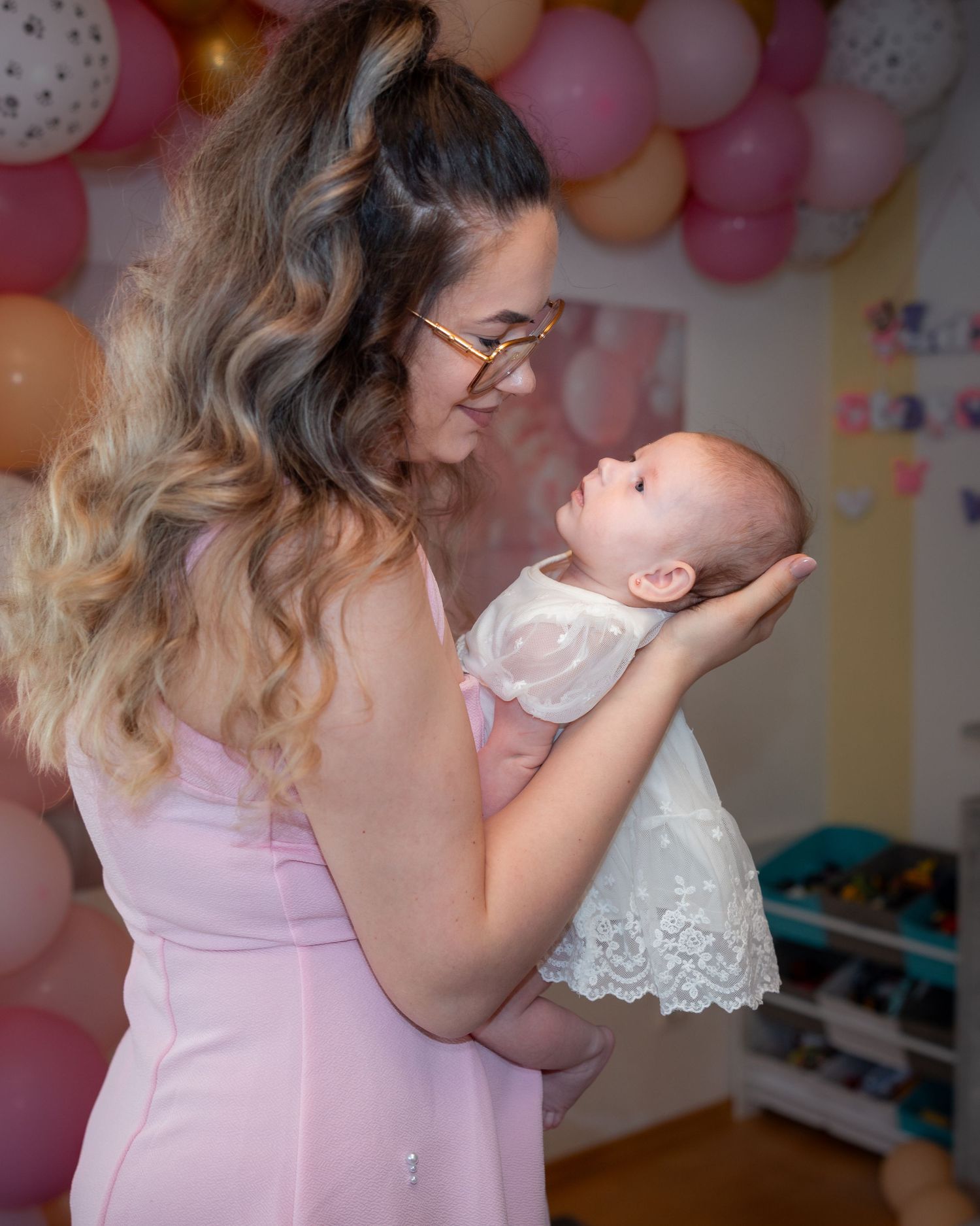 Eine Mutter im rosa Kleid hält ihr Baby behutsam und blickt ihm vor einer Ballondekoration in die Augen.