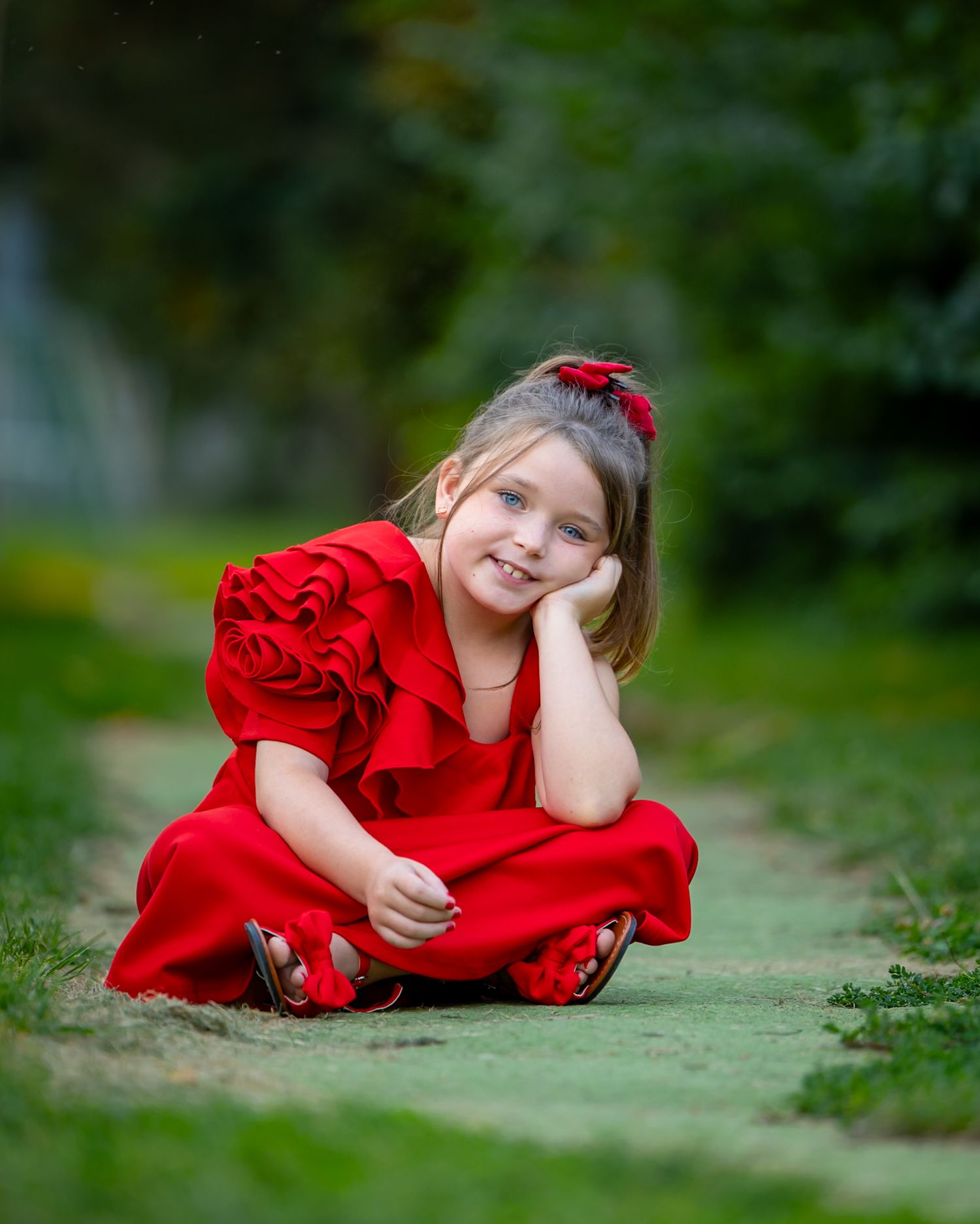 A young girl in a red dress and hair bow sits cross-legged on a garden path, smiling with her chin resting on her hand.