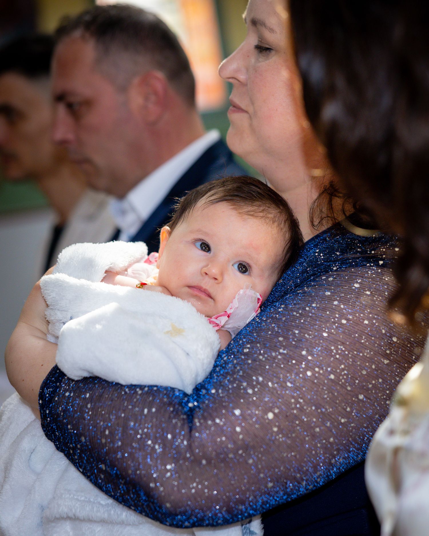 A woman in a blue glittery dress tenderly holds a newborn baby at what appears to be a christening ceremony.