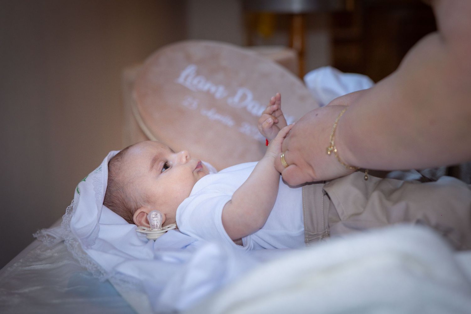Newborn baby lying on a bed in a white onesie with a pacifier, gently holding an adult's hand.