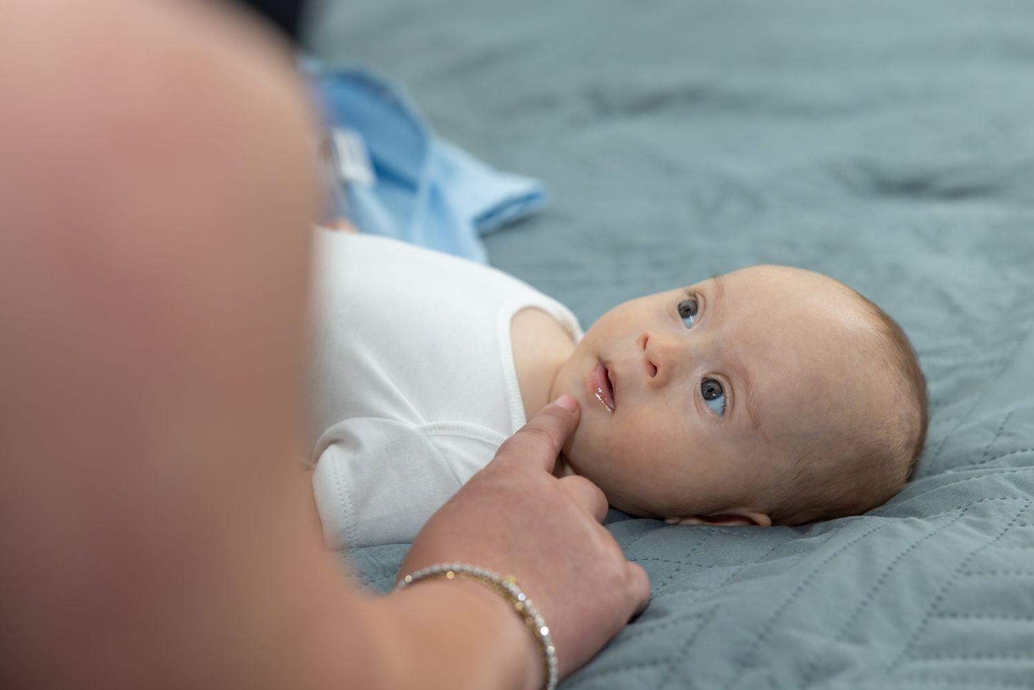 A newborn baby in a white onesie lies on a blue blanket while an adult gently touches their chin.