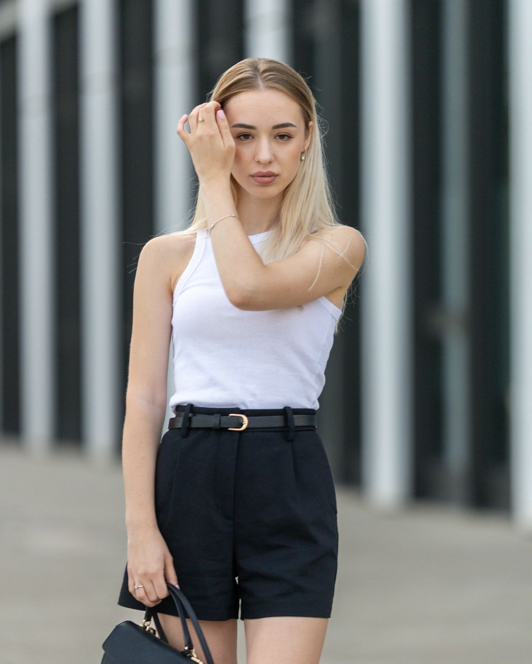 Young blonde woman in white tank top and black belted shorts holding a handbag outdoors.