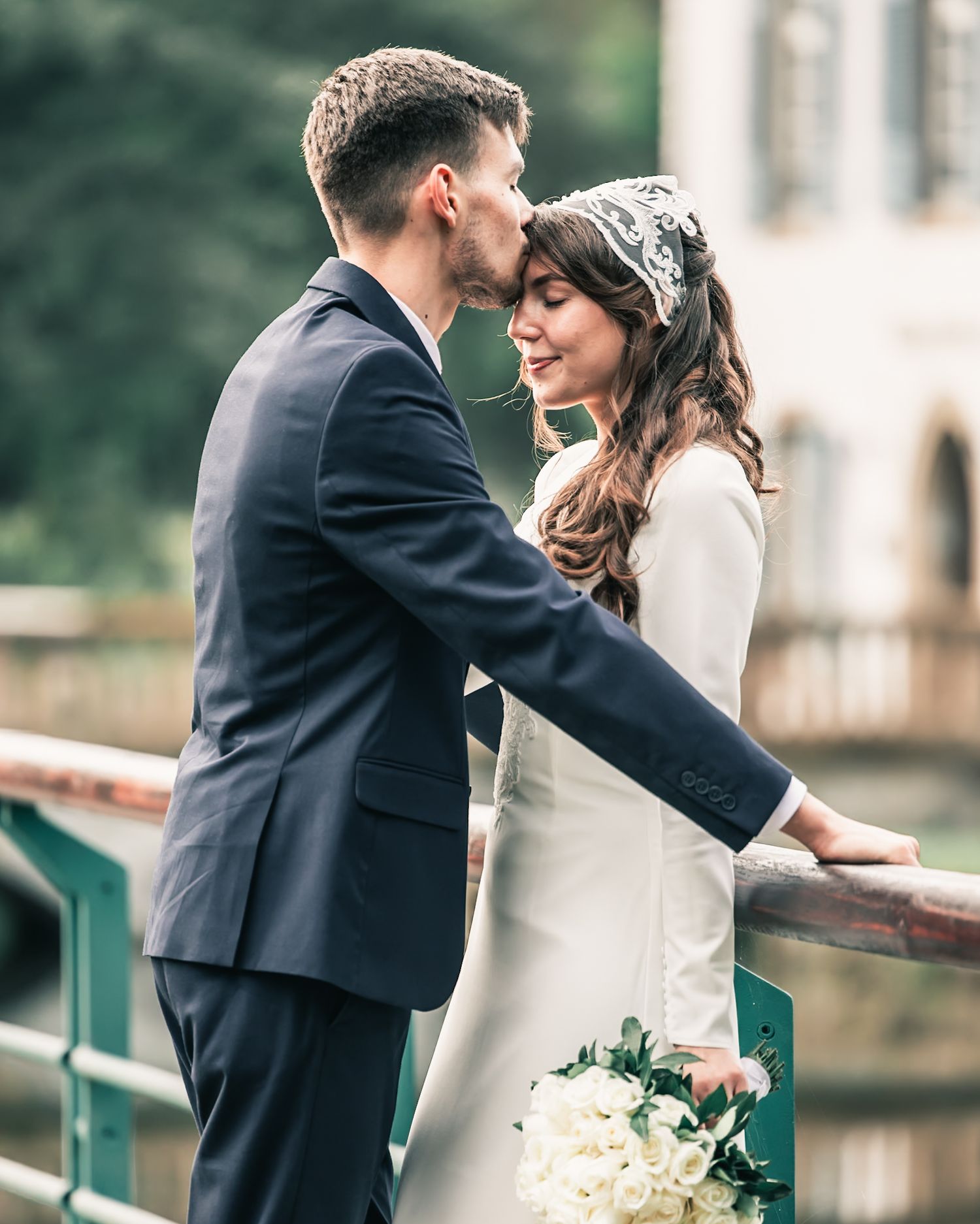 Bride and groom share a romantic kiss on a bridge, bride holding white bouquet in elegant white dress and lace headpiece.
