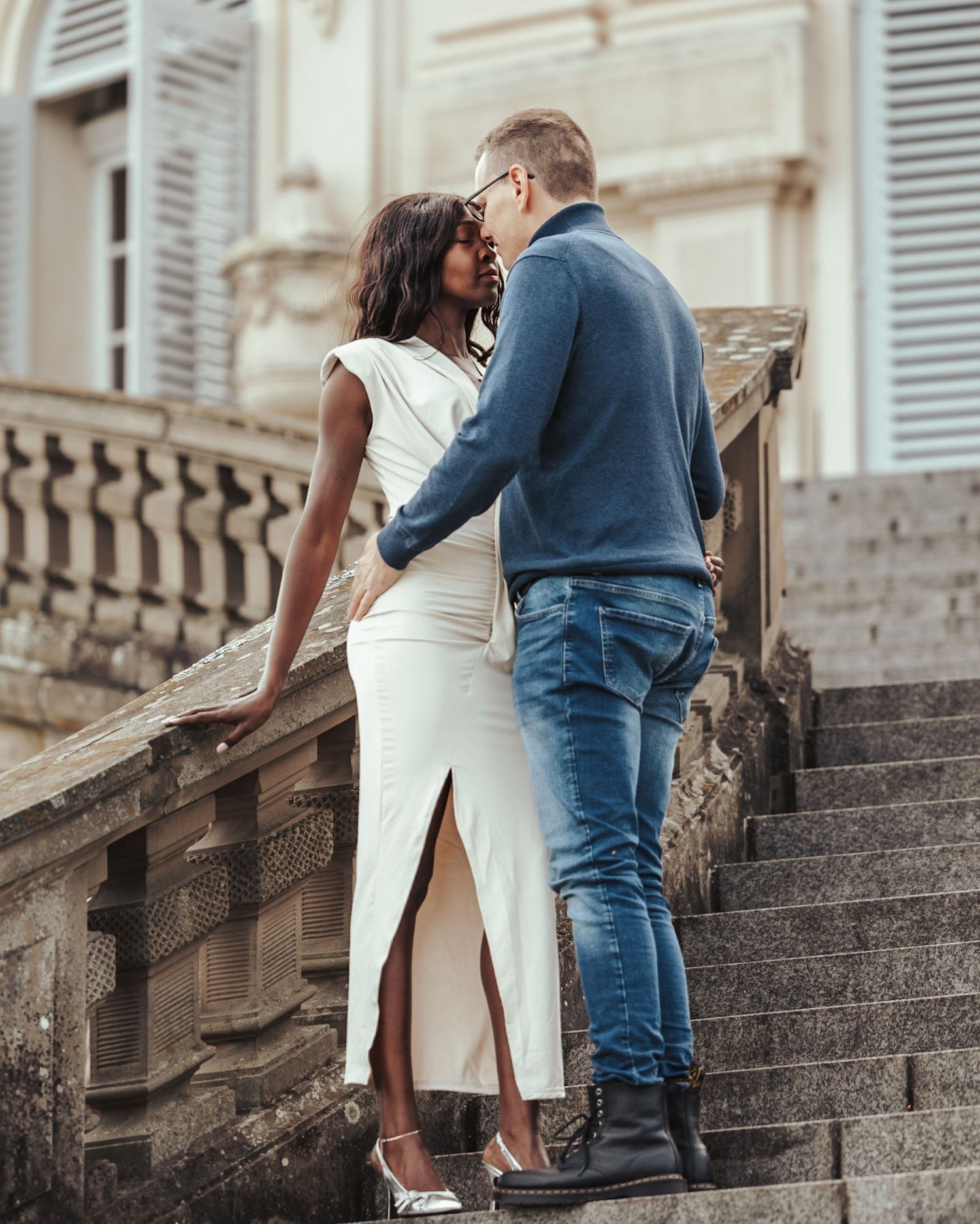 Couple embracing on stone staircase in front of elegant building with white dress and blue denim outfit.