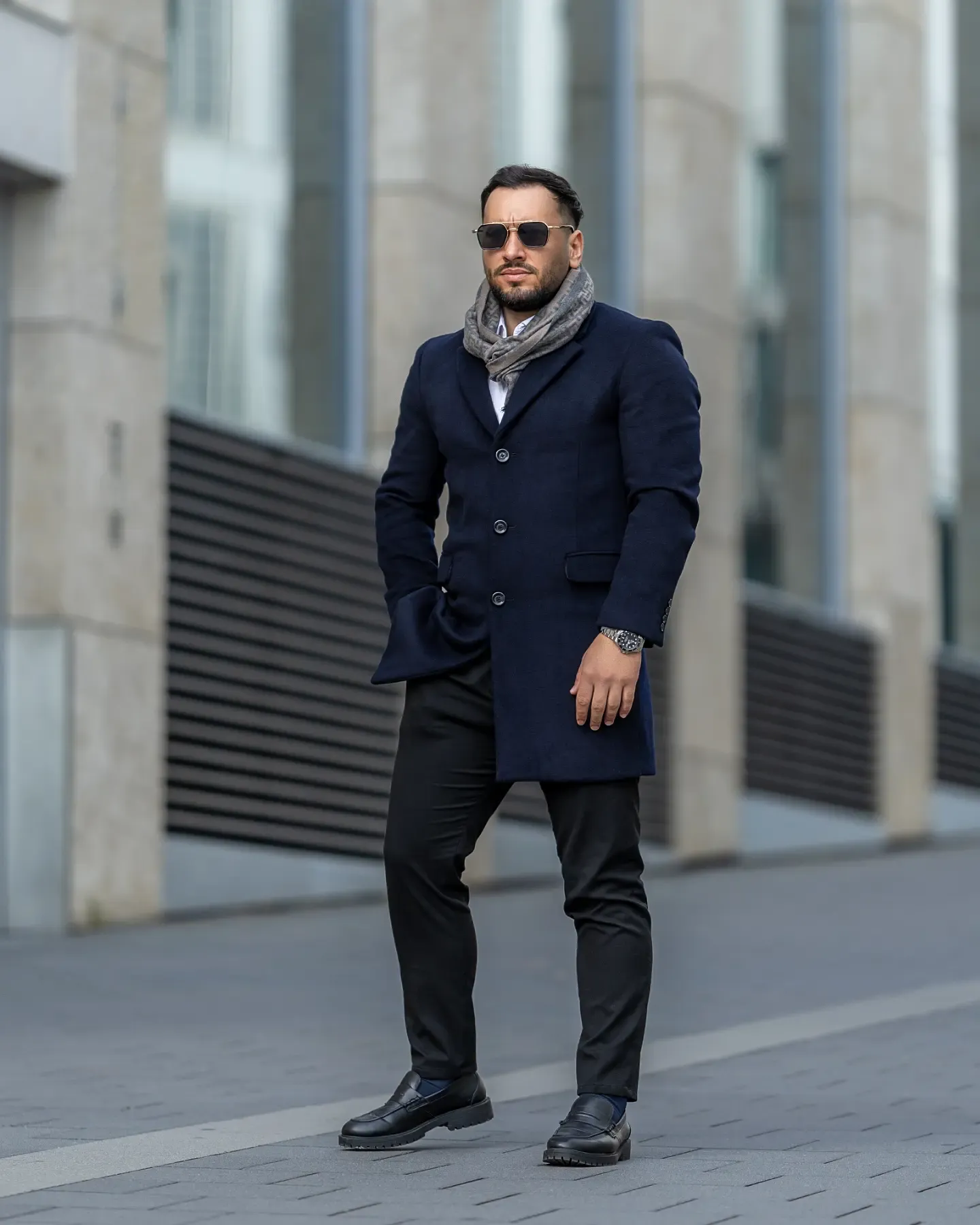 Man in navy blue coat, gray scarf, black pants, and leather shoes posing outdoors near a modern building.