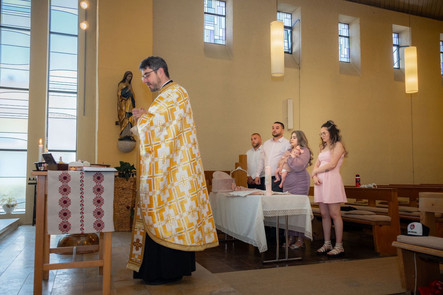 Der Priester führt die Taufzeremonie; Lara wird hinter ihm im Kreis ihrer Familie auf dem Arm gehalten.