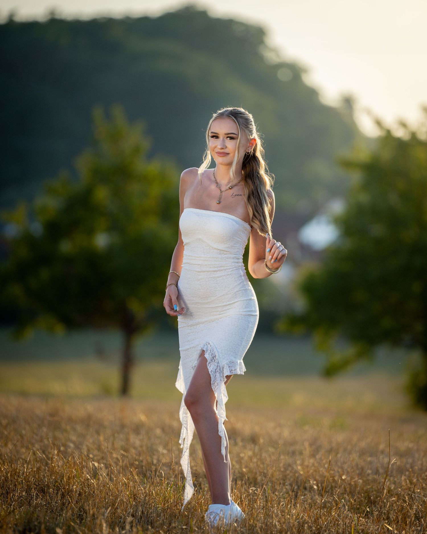 Ganzkörperporträt von Cătălina im weißen Kleid zwischen trockenen Gräsern, mit Bäumen und hellem Himmel im Hintergrund.
