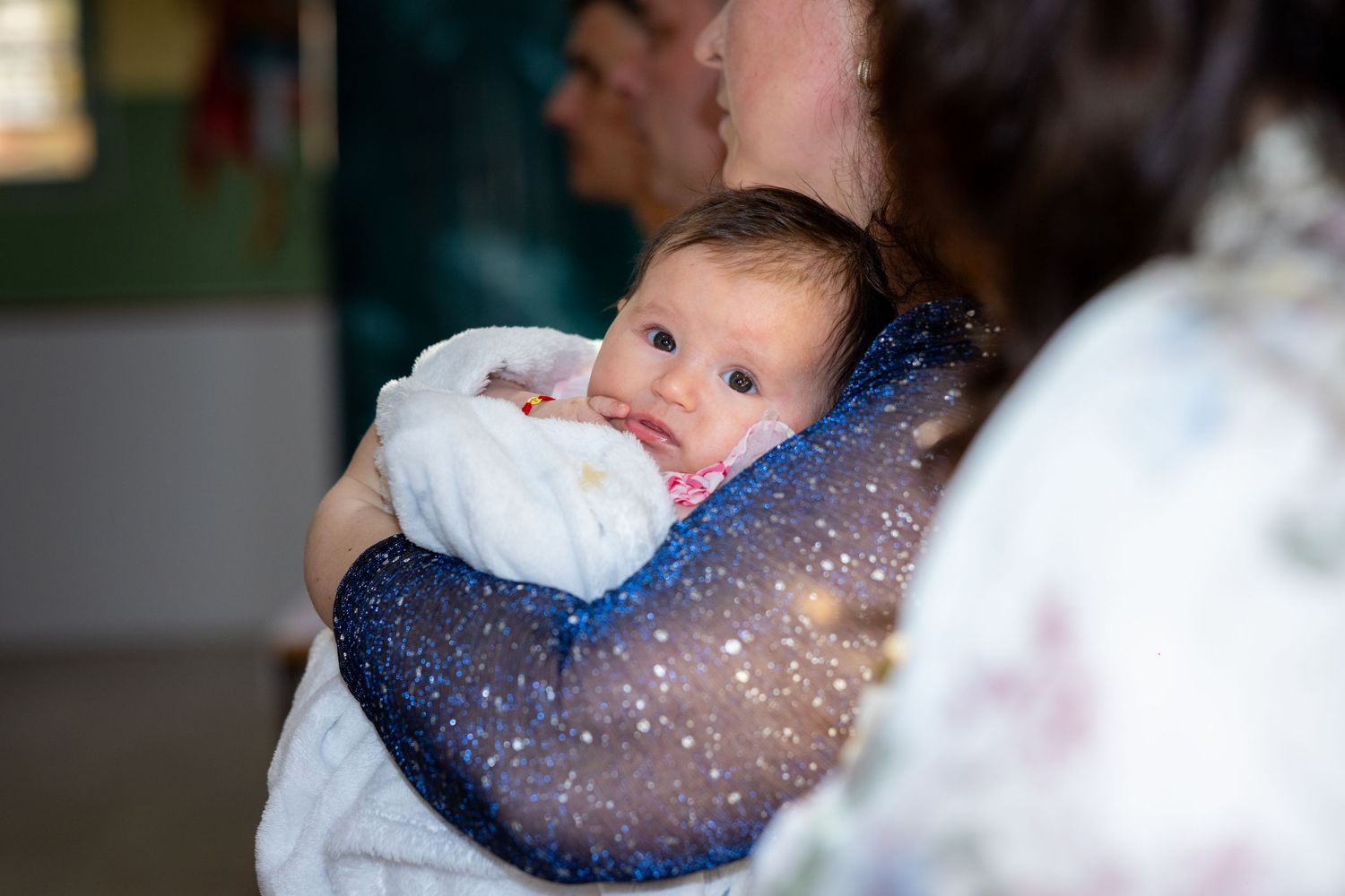 A newborn baby wrapped in a white blanket being held by a person in a blue sparkly dress at a christening.