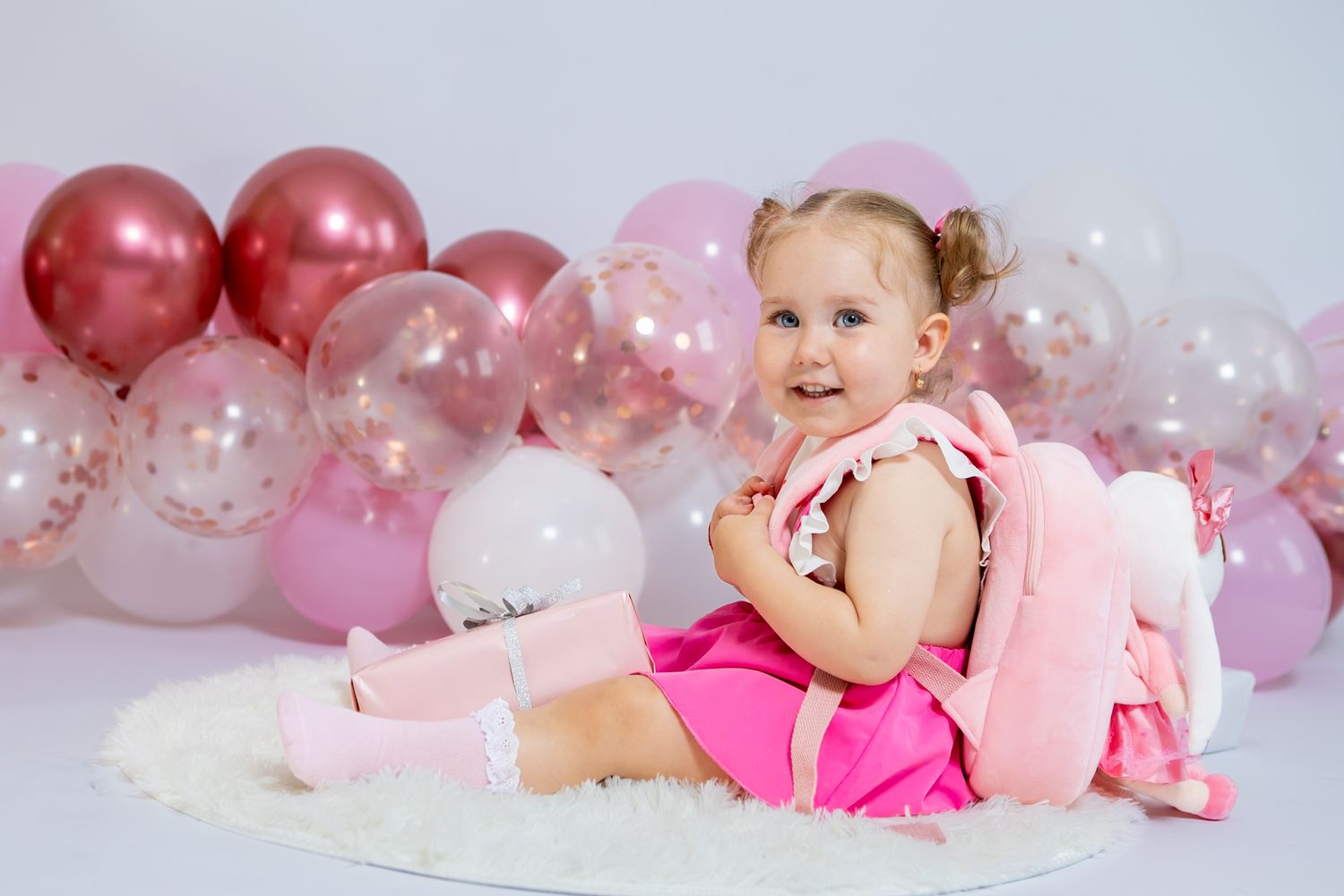 Cute baby girl in pink tutu sitting on white fur rug surrounded by pink and rose gold balloons at birthday party.