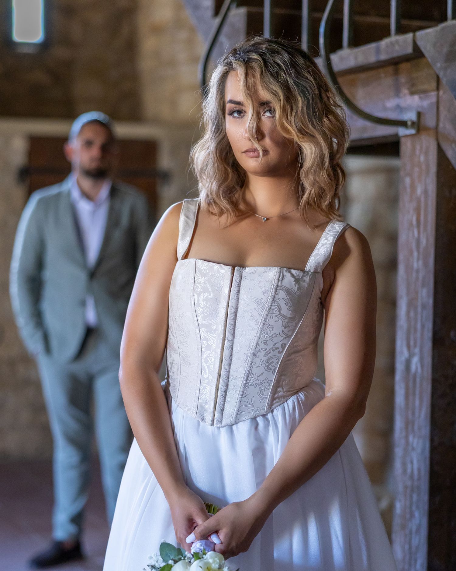 Woman in white wedding dress holding flowers with man in suit standing in background in rustic indoor setting.