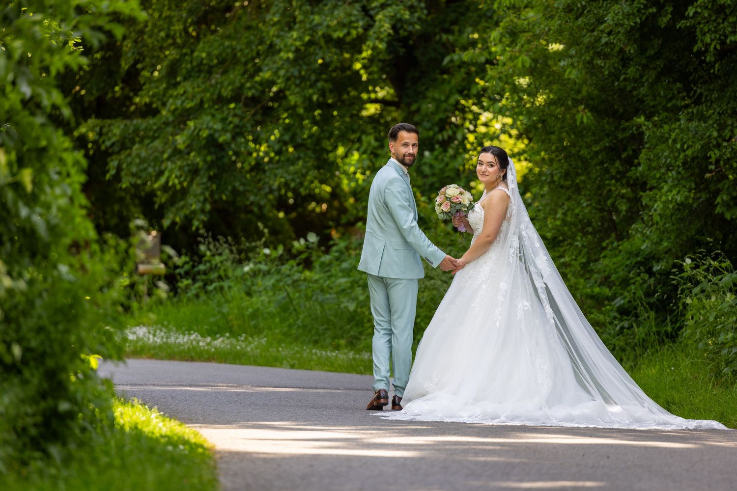 Angeliki und Dominik stehen Hand in Hand auf einem Weg zwischen grünen Bäumen.