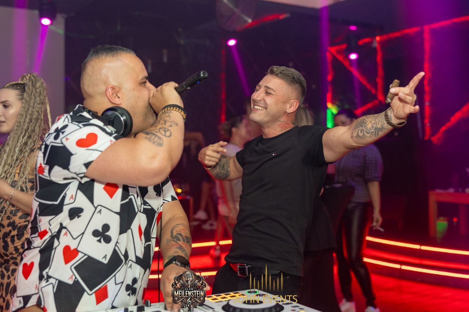 Ein Mann singt ins Mikrofon, während ein anderer neben dem DJ-Pult im Club Meilenstein die Arme hebt.
