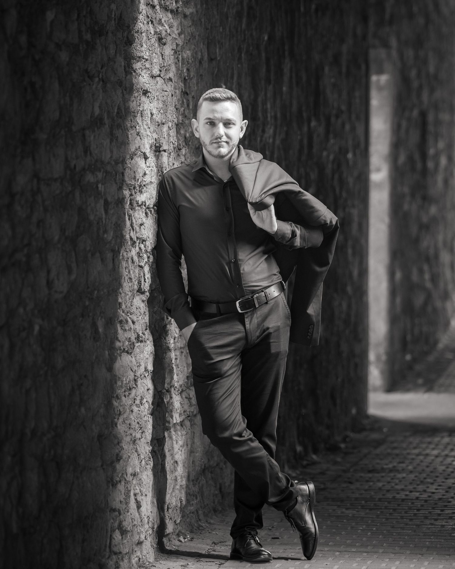Schwarz-Weiß-Portrait eines Mannes mit dunklem Hemd und Jacke über der Schulter, der an einer Steinwand lehnt.
