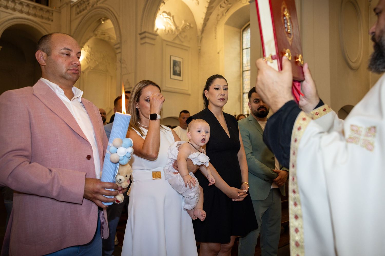 Der Priester hält ein rotes Buch vor der Taufgesellschaft; Aris wird neben der brennenden Kerze getragen.