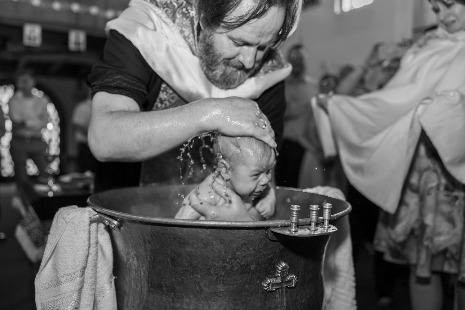 Black and white photo of a bearded priest baptizing a baby in a traditional metal font during a church ceremony.