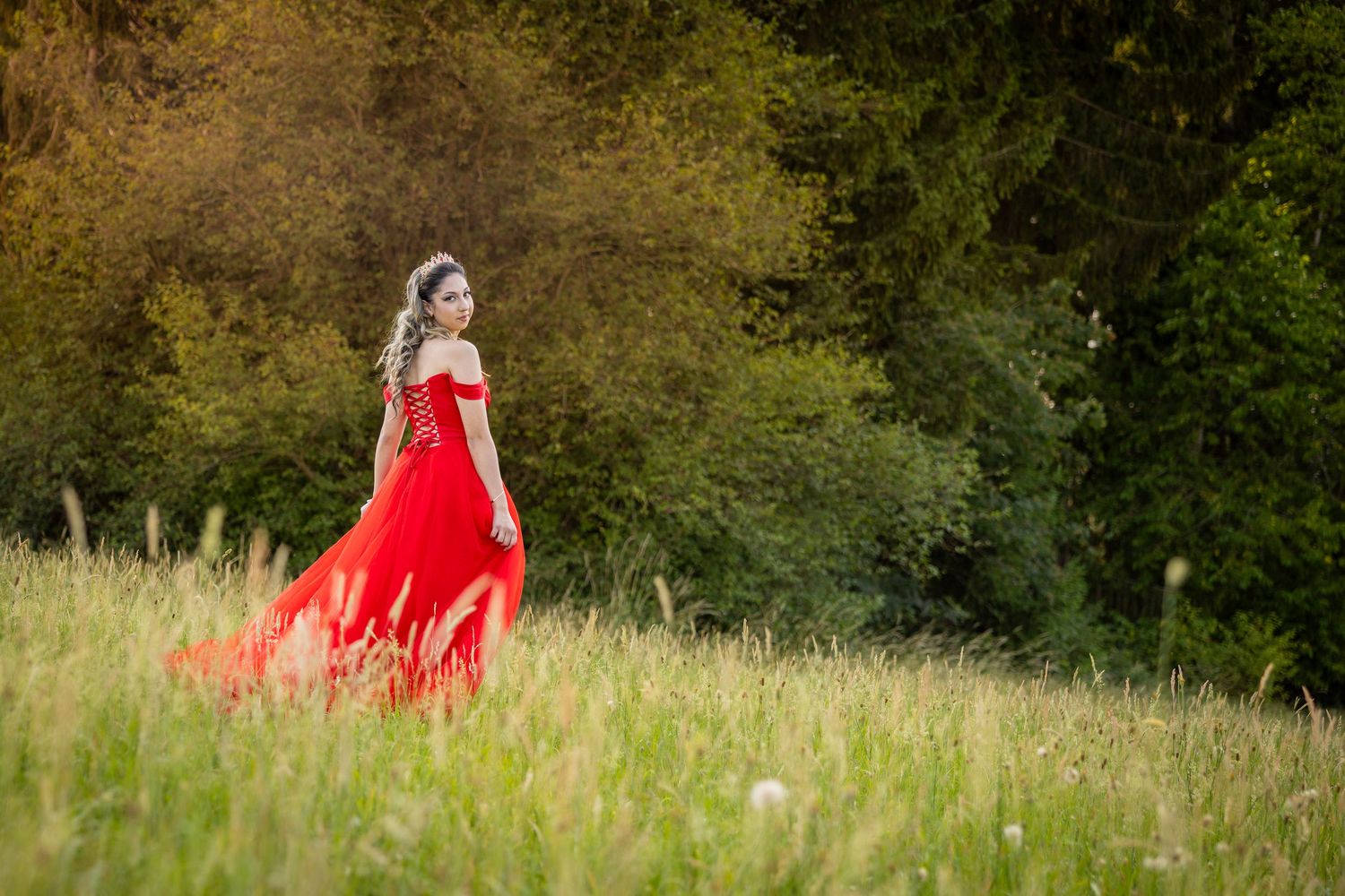 Junge Frau im langen roten Kleid geht durch hohes Gras und blickt über ihre Schulter zur Kamera.