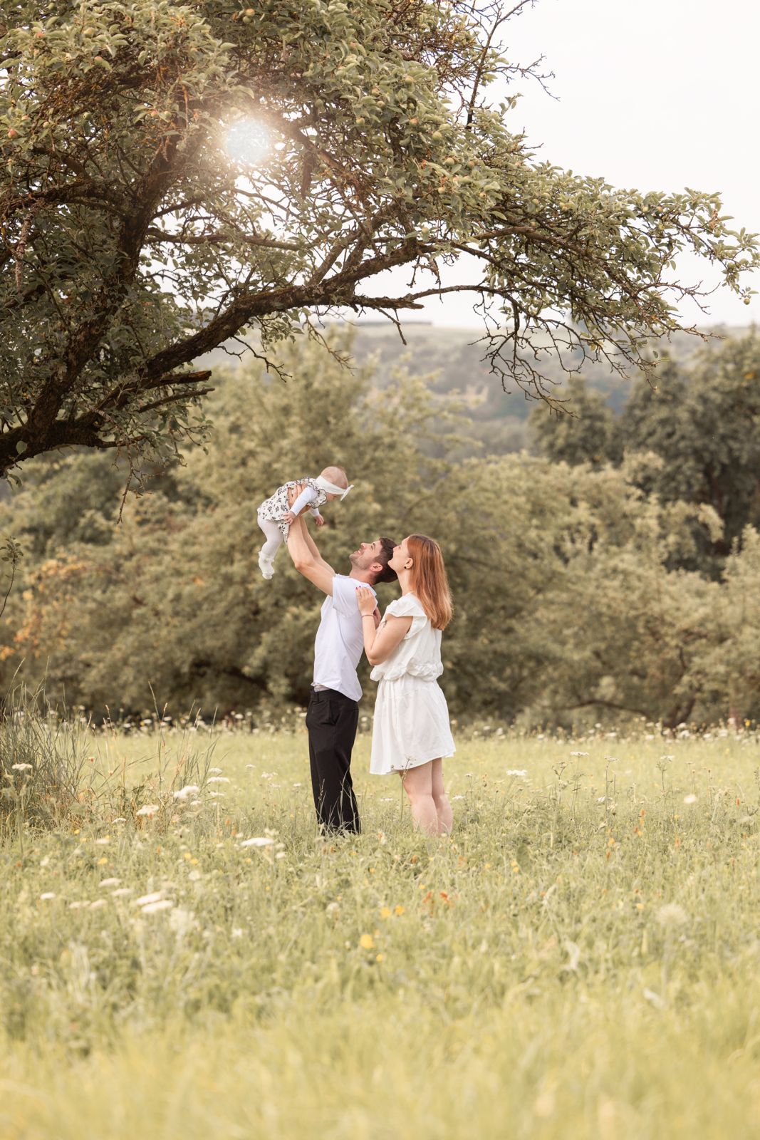 Happy young family with baby enjoying sunny day in green meadow, father lifting child while mother watches lovingly.