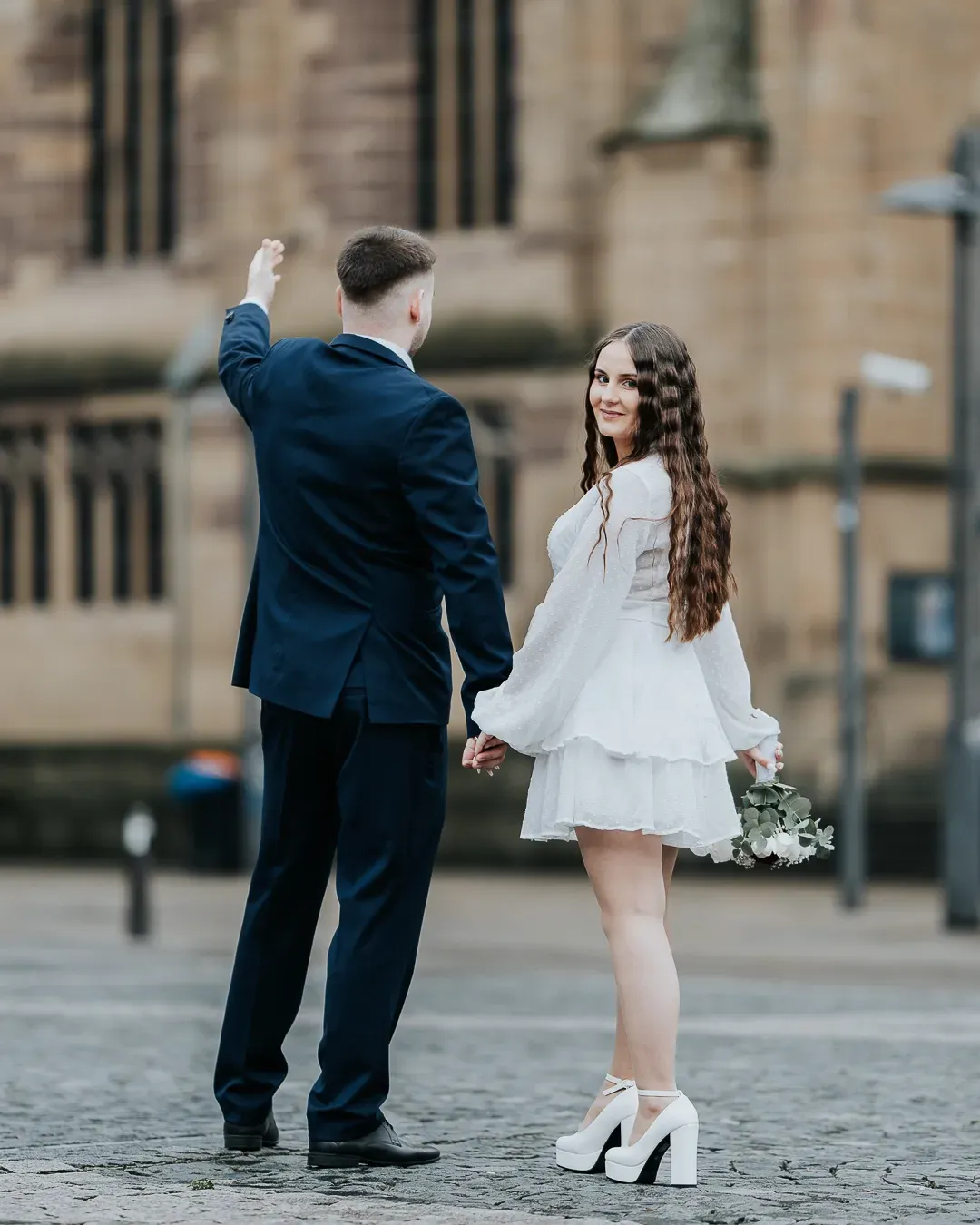 Bride in white mini dress and platform heels holds flowers while groom in navy suit spins her on cobblestone plaza.