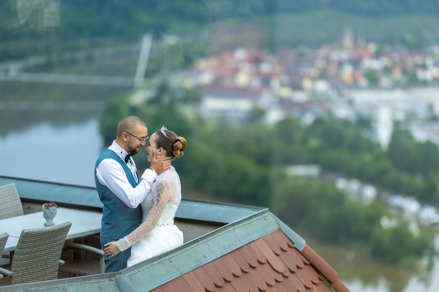 Bride and groom embracing on a rooftop with a scenic river town view in the background.